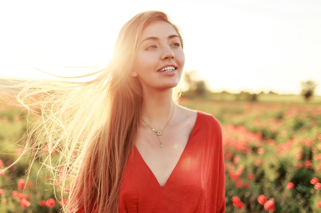 Нежная белокурая длинноволосая женщина с идеальной улыбкой позирует на маковом поле в теплый летний закат.