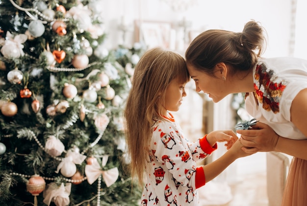 Нежная мама с маленькой дочкой в пижаме у новогодней елки в светлой уютной комнате.