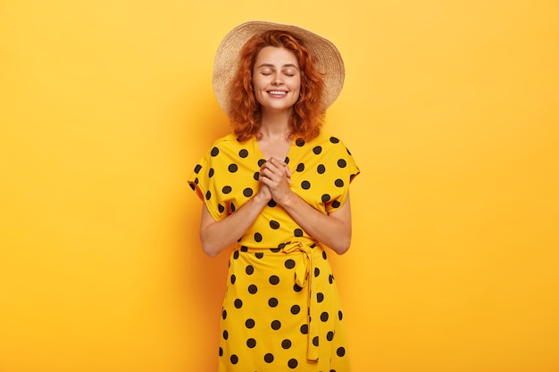 Gentle pleased redhead woman posing in yellow polka dress and straw hat