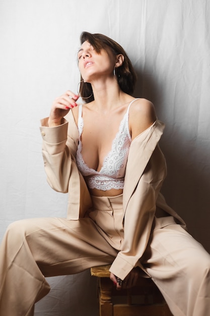 Free Photo  Gentle portrait of a beautiful young woman with big breasts in  a beige suit and white lace bra sits on a chair on a gray white
