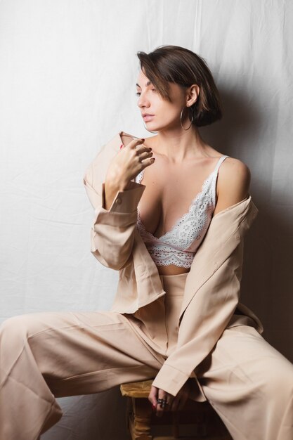 Gentle portrait of a beautiful young woman with big breasts in a beige suit and white lace bra sits on a chair on a gray white