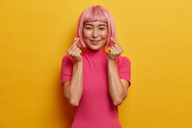 Нежная улыбающаяся симпатичная женщина с модной розовой стрижкой, жестикулирует по-корейски, выражает любовь, находится в хорошем настроении.
