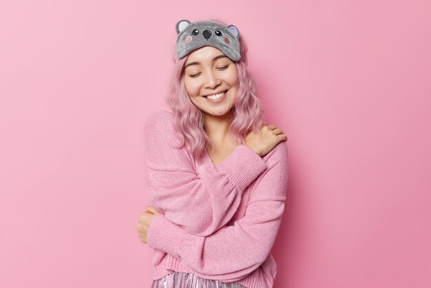 Gentle smiling woman with dyed hair embraces herself and daydreaming keeps eyes closed wears sleepmask and soft jumper recalls nice moment in life isolated over pink background. Self care concept