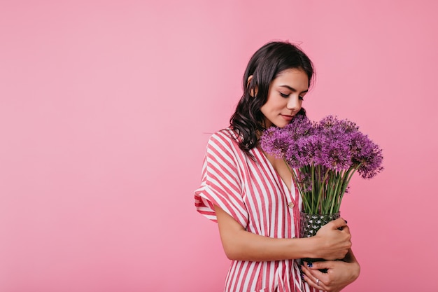 Gentle young woman in romantic mood is cute looking at armful of flowers. Portrait of European lady in stylish outfit.