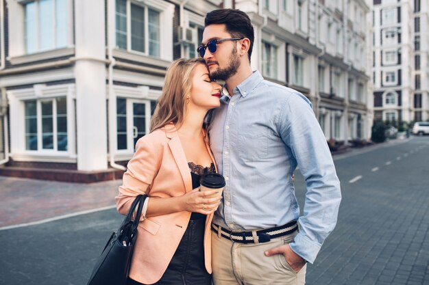 Gently couple is hugging on street in city. Handsome guy  is blue shirt and sunglasses looks seriously, pretty blonde girl in black dress cuddling to him.