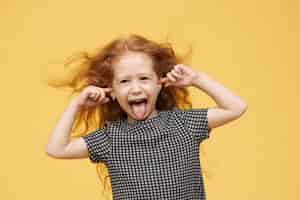 Free photo genuine human emotions, reaction and body language. angry spoiled little girl with red hair sticking out tongue, pretending not to hear you, plugging ears, screaming, being mad and naughty