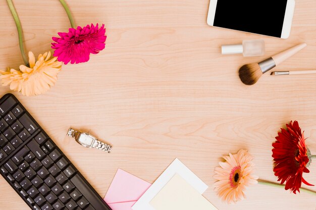 Gerbera flowers; keyboard; wrist watch; envelope; makeup brush; nail polish bottle and cellphone on wooden desk