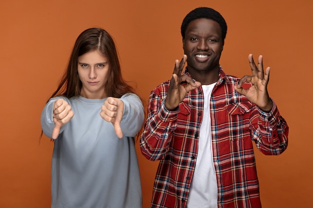 Gestures, symbols and signs concept. Emotional interracial couple expressing controversial attitude - black man smiling and making ok gesture while annoyed angry white woman showing thumbs down