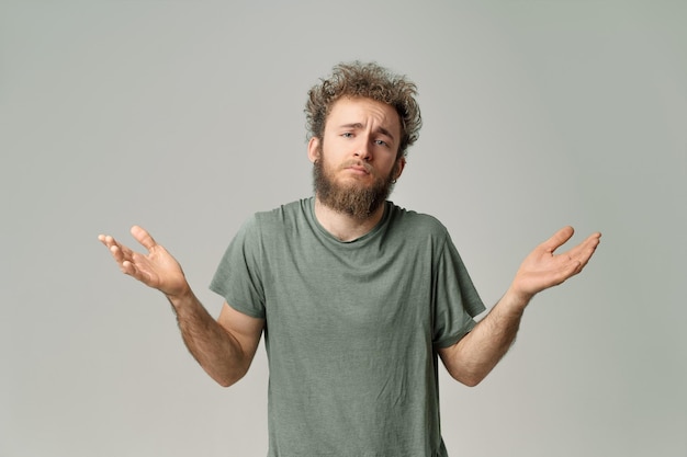 Gesturing I DONT KNOW or I AM SORRY young handsome bearded wild curly hair man with bright blue eyes isolated on grey background Young thinking man in green t shirt on white Copy space