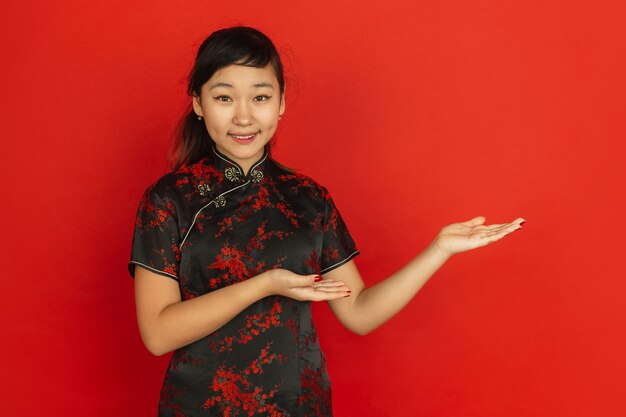 Gesturing, inviting guests. Happy Chinese New Year 2020. Asian young girl's portrait on red background. Female model in traditional clothes looks happy. Celebration, human emotions. Copyspace.