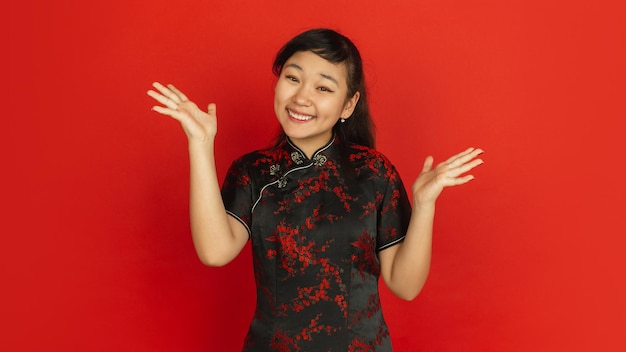 Жесты, улыбка, приглашение. Счастливого китайского Нового года. Портрет азиатской молодой девушки на красном фоне. Женская модель в традиционной одежде выглядит счастливой. Праздник, человеческие эмоции. Copyspace.