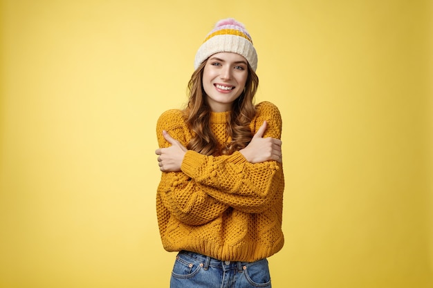 Getting cold bring jacket. Portrait charming tender feminine cute girlfriend embacing herself cuddling smiling warm gaze freezing outside standing knitted sweater hat, yellow background