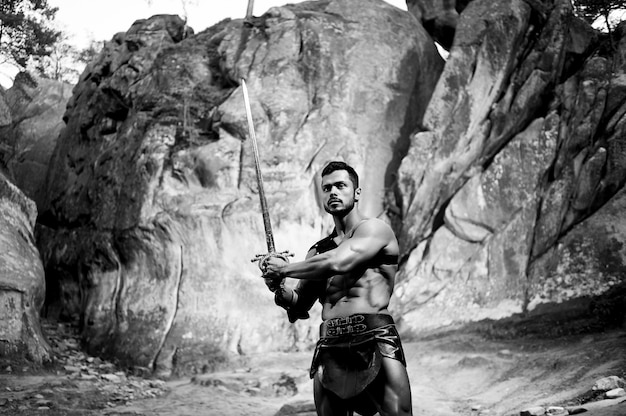 그의 전투를 준비합니다. 바위 근처에서 용감하게 포즈를 취하는 칼을 든 용감한 남자 전사의 흑백 초상화
