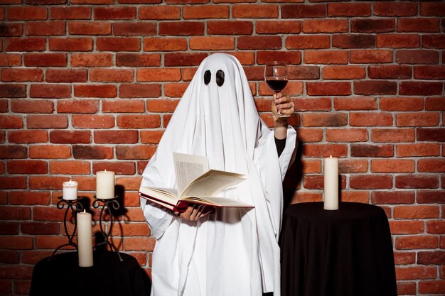 Ghost holding book and wine over brick wall. Halloween party.