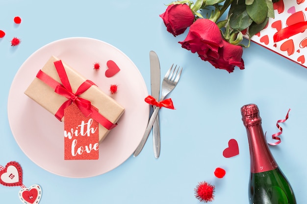 Gift with label on plate near cutlery, roses and bottle of champagne