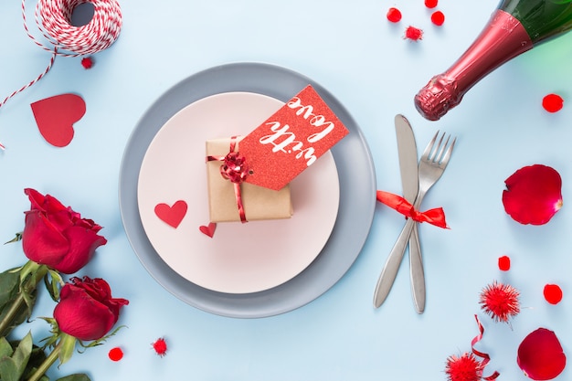 Gift with tag on plate near cutlery, roses and bottle of champagne