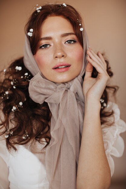 Ginger curly woman in beige headscarf and flowers in hair gently looks at camera.