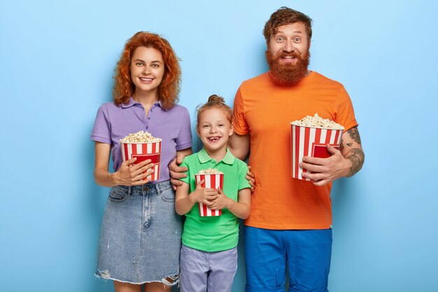 Ginger family spend free time in cinema, watch funny movie, smile happily, eat delicious popcorn, stand closely to each other, enjoy togetherness, entertain themselves. Leisure, family time concept