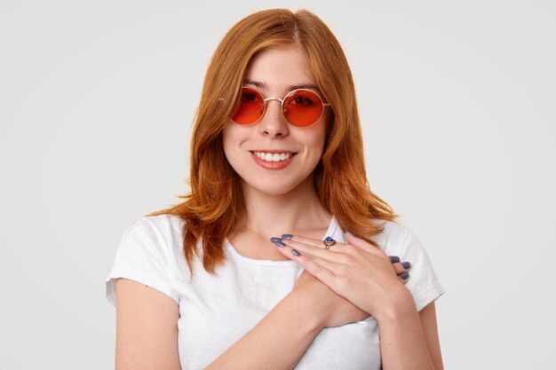 Ginger female with positive smile, keeps hands on chest, expresses good attitude, wears trendy red shades