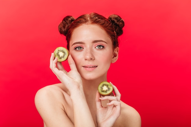 Ginger girl holding kiwi. Studio shot of caucasian adorable woman with tropical fruits.