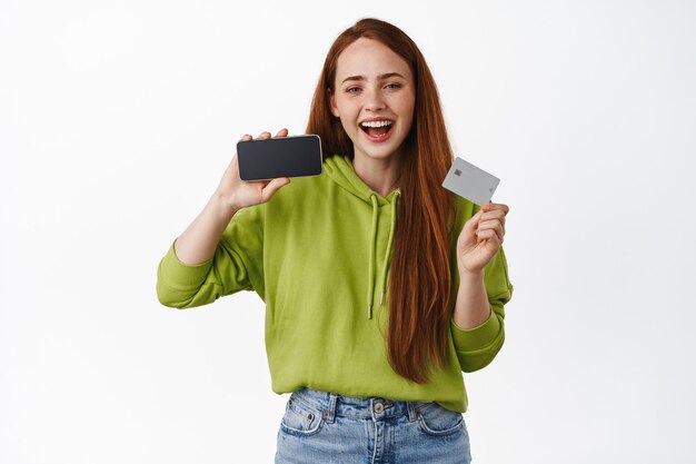 ginger girl makes purchased, paying in internet, showing credit card and horizontal smartphone screen on white