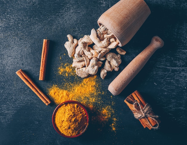 Ginger powder in a bowl with herb grinder, ginger and dry cinnamon pack top view on a dark textured background