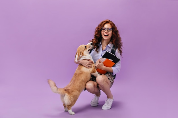 Ginger woman in blue shirt and eyeglasses playing with corgi and holding notebooks Smiling girl with wavy hair posing with fanny dog