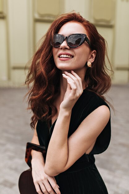 Рыжая женщина в модных солнцезащитных очках позирует на улице и улыбается