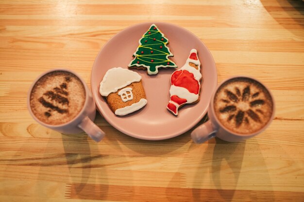 クリスマスデザインのジンジャーブレッドは、コーヒー入りカップの間にピンクのプレートの上に横たわっています
