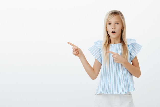 Girl amazed seeing magician trick. Portrait of fascinated admiring young blond daughter in trendy blue blouse, saying wow, pointing left with index finger, feeling astonished, gasping over gray wall