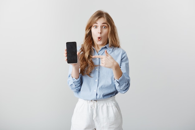 Girl amazed with new phone. portrait of fascinated shocked young european woman with blond hair in blouse showing smartphone, pointing at device, saying wow, expressing astonishment