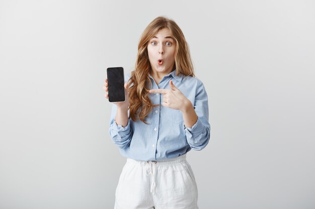 Девушка поразила новым телефоном. Портрет очарованной шокированной молодой европейской женщины со светлыми волосами в блузке, показывающей смартфон, указывающей на устройство, говорящей вау, выражающей удивление