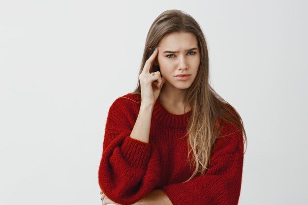 Girl annoyed, do not want continue conversation. Bothered displeased caucasian female entrepreneur in loose sweater, holding index finger on temple, frowning, doubtful and confused over gray wall