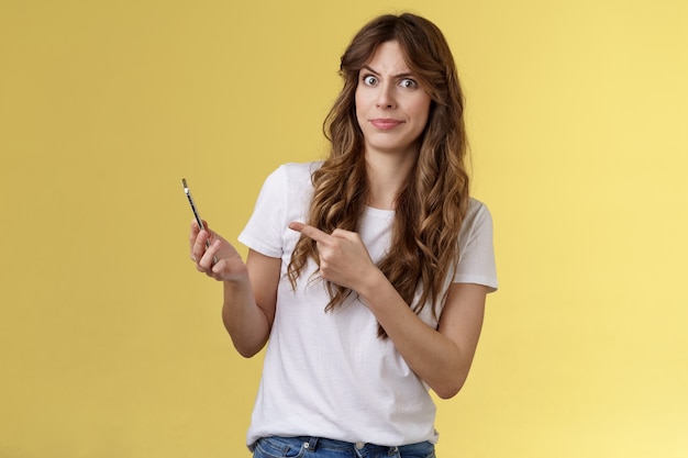 Girl answers strange weird call receive crazy upsetting message cringe doubtful displeased smirking dismay pointing suspicious smartphone stand yellow background intense frustrated