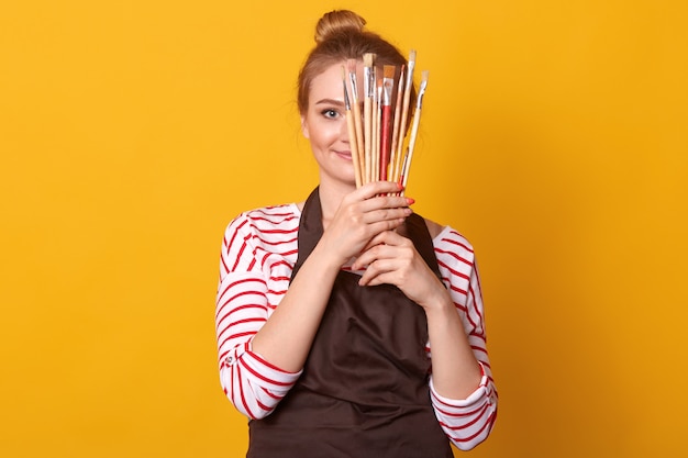 Girl artist holds brushes in hands and hides behind it, lady dresses stripes casual shirt and brown apron, blonde woman with bunch