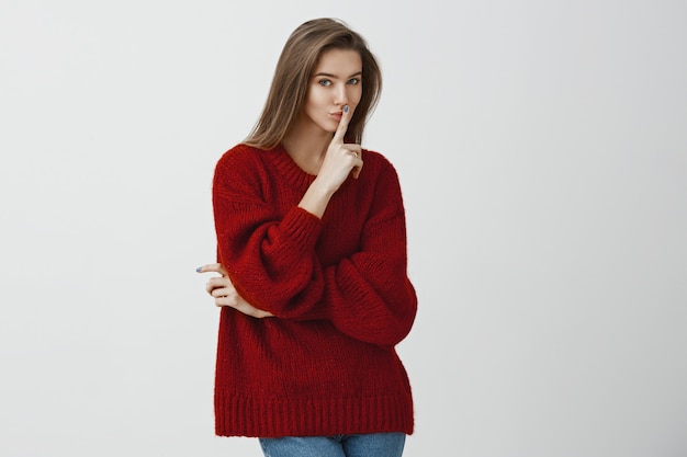 友達に秘密を守るように頼んで誰にも言わなかった女の子。スタイリッシュなルーズな赤いセーターで遊び心のある軽薄なヨーロッパの女性、人差し指を口にかざし、興味をそそる笑顔、shhまたはshush記号を作る