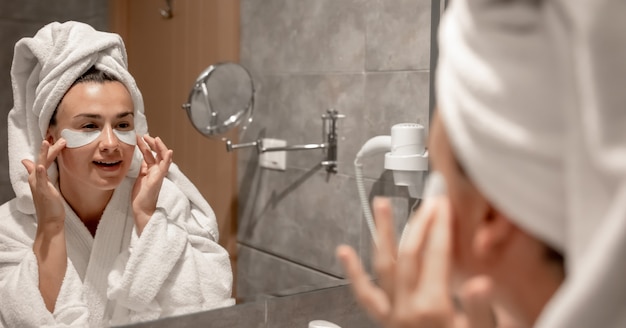 A girl a bathrobe and with a towel on her head sticks patches under her eyes in the bathroom in front of the mirror.
