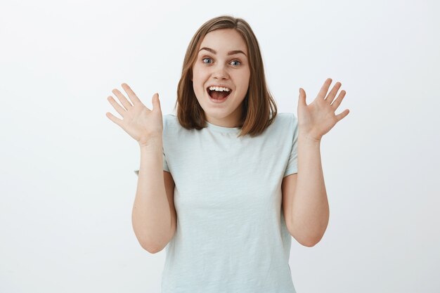 여자 친구가 예기치 않게 어깨 근처에 손바닥을 들고 회색 벽 위에 트렌디 한 티셔츠에 서있는 놀라움과 긍정적 인 감정에서 즐겁게 웃고있는 것을보고 흥분하고 놀란 소녀