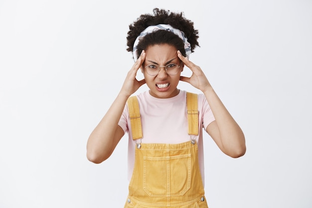 Free photo girl believes she can move object with power of mind. portrait of funny and angry cute female in dungarees, headband and glasses, frowning and clenching teeth with anger, holding fingers on forehead