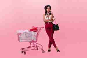 Free photo girl in beret and bright trousers looks at camera thoughtfully and poses next to supermarket trolley. photo of woman in stylish bright outfit on pink background.