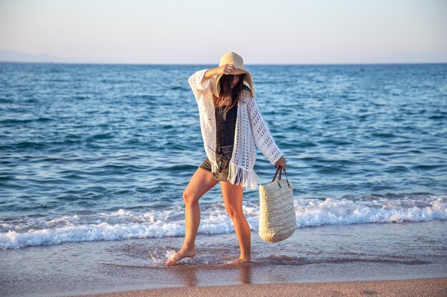 籐のバッグを持った大きな帽子をかぶった女の子が海岸を歩きます。夏休みのコンセプト。