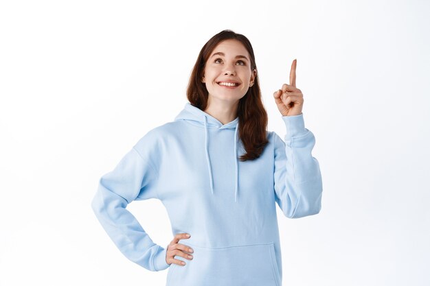 Girl in blue hoodie pointing and looking up at promo logo, smiling pleased, showing good advertisement, standing against white wall