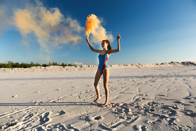 Girl in blue swim-suit dances with orange smoke on white beach