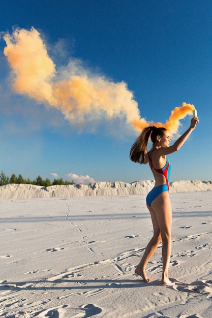 Girl in blue swim-suit dances with orange smoke on white beach