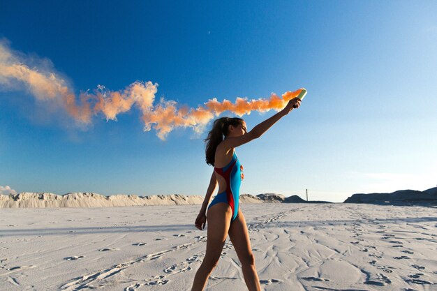 Girl in blue swim-suit dances with orange smoke on white beach