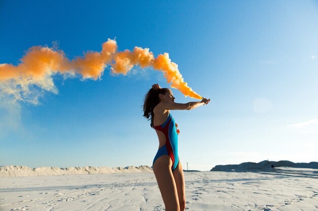 Girl in blue swim-suit dances with orange smoke on white beach