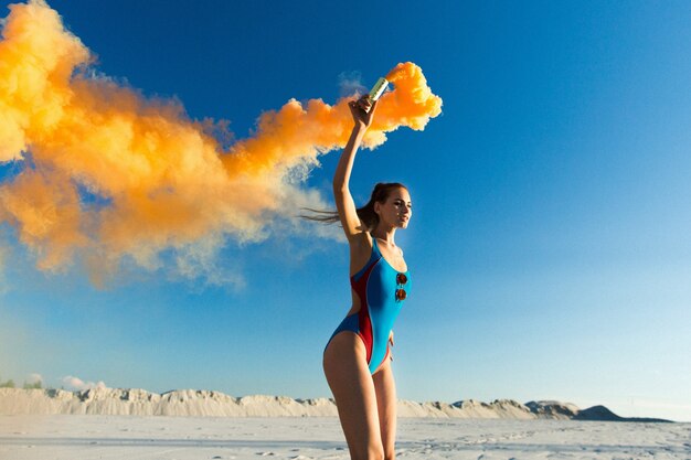 Girl in blue swim-suit dances with orange smoke on white beach