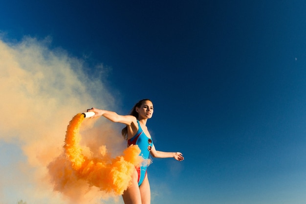 Girl in blue swim-suit dances with orange smoke on white beach