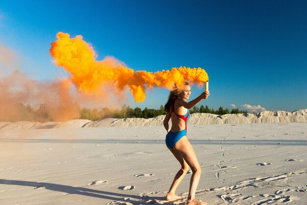 Girl in blue swim-suit dances with orange smoke on white beach