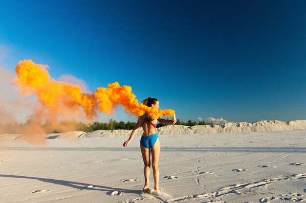 Girl in blue swim-suit dances with orange smoke on white beach